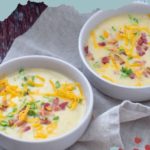 Two bowls of delicious baked potato soup, brimming with flavor, are topped with shredded cheese, crispy bacon bits, and fresh green onions, all beautifully arranged on a cloth.