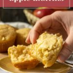 A hand holding a partially cut open muffin with butter spread, placed on a yellow plate. Apple muffins and fresh apples are in the background. Text overlay reads, "Maple apple muffins.