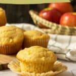 Several apple muffins displayed on a wooden surface, with some muffins in the foreground and a basket of red apples in the background. The text reads "maple apple muffins, tender & delicious.