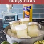 Mason jars brimming with tangy margaritas nestle in an ice-filled metal tub, flanked by a bottle of tequila, orange liqueur, zesty limes, and a vibrant green cloth on the dark countertop.