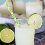 A mason jar brimming with margaritas, garnished with a lime slice and a green straw, sits invitingly in front of a metal tub filled with ice.