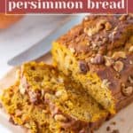 A loaf of delicious pumpkin persimmon bread topped with chopped nuts is partially sliced on a wooden board, with a knife resting in the background.