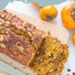 A loaf of pumpkin persimmon bread with walnuts is sliced on a wooden cutting board. Near the bread are whole persimmons. The image has a text overlay that reads, "Pumpkin Persimmon Quick Bread.