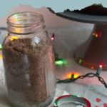 A jar of homemade devil's food cake mix sits invitingly on a table, its lid ajar while colorful string lights twinkle warmly in the background.