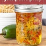 A jar of tangy jalapeño jelly sits on a wooden surface, with a fresh jalapeño pepper and a bowl of crackers in the background.