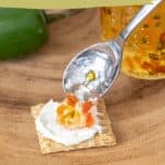 A spoon of jalapeño jelly with pieces of jalapeño is being placed on a cracker with cream cheese. In the background, a jar of jalapeño jelly and a whole jalapeño pepper complete the appetizing scene.