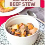 A bowl of classic beef stew, brimming with hearty chunks of beef, potatoes, and carrots, rests on a patterned placemat. In the background, a fork lies beside a plate ready to receive its share of rich flavor and warmth.