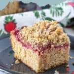 A slice of cranberry coffee cake with pecan streusel sits on a dark plate, featuring a crumbly topping and a layer of cranberry filling. The background includes festive decorations.