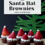 Festive santa hat brownies adorned with strawberries and whipped cream are artfully arranged on a wooden tray against a vibrant green backdrop.