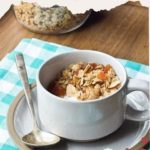 A mug brimming with homemade granola and apricots rests on a saucer, complete with a spoon. Beneath it, a blue and white checkered cloth adds charm. Text reads, "Homemade Granola with Apricots.