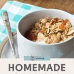 A white bowl brimming with homemade granola and apricot pieces rests on a pristine plate. Beside it, a silver spoon lies on a checkered napkin, while a jar of the crunchy delight waits eagerly in the background.