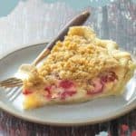 A luscious slice of raspberry cream pie with streusel topping is elegantly served on a white plate, accompanied by a fork ready to indulge you.