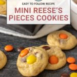 A close-up of Mini Reese's Pieces cookies on a wooden surface, showcasing the vibrant colors and delightful texture, with more delectable Reese's Pieces cookies tempting from a tray in the background.