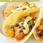 Three delicious butternut squash tacos with black beans, creamy avocado, and a fresh garnish sit invitingly on a plate. Text overlay reads "butternut squash tacos" and "vegetarian recipe," with the website URL subtly displayed.