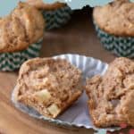 A close-up of apple cinnamon muffins, with one muffin cut in half to reveal the inside. The delectable treats are nestled in green and white striped paper liners. Text at the top reads "APPLE CINNAMON muffins.