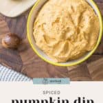 A bowl of pumpkin cream cheese dip is displayed on a wooden surface next to apple slices and a whole nutmeg seed, adding an aromatic touch.