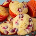 A tray filled with cranberry orange muffins in vibrant orange wrappers, showcasing a light, fluffy texture with visible cranberry pieces. Above, the text proudly declares "Cranberry Orange Muffins.