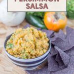 A vibrant bowl of persimmon salsa sits ready to be enjoyed with crisp tortilla chips, surrounded by its fresh ingredients: a whole onion, spicy jalapeño, and ripe persimmon.