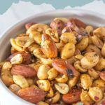 A vibrant bowl of furikake roasted almonds and peanuts sits against a blue backdrop, adorned with decorative text and intricate patterns.