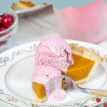 A plate holds a slice of pumpkin pie adorned with a scoop of cranberry ice cream, its pink hue adding a vibrant touch. A fork rests invitingly on the plate.