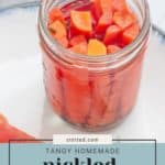 A glass jar filled with sliced, tangy homemade pickled carrots is labeled "Tangy Homemade Pickled Carrots" with a website name above, inviting you to discover how to pickle carrots yourself.