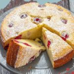 A lemon raspberry ricotta cake sits elegantly on a grey cake stand, sliced into perfect pieces. A light dusting of powdered sugar graces the top, enhancing its delightful simplicity.