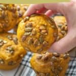 Hand holding a pumpkin muffin with pepitas, topped with spiced seeds, surrounded by more muffins on a checked cloth, with text "pumpkin muffins with spiced pepitas" and a website link.