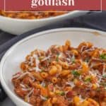 A steaming bowl of classic American goulash features macaroni, savory ground meat, and rich tomato sauce, crowned with a sprinkle of shredded cheese and fresh herbs.