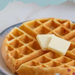 A round buttermilk waffle with two pats of butter sits invitingly on a plate, proudly displaying the text "Buttermilk Waffles," with a website name subtly listed below.