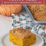 A slice of pumpkin coffee cake with pecan cinnamon streusel sits on a plate, placed in front of a tray of the fresh-baked cake. A decorative pumpkin and autumn-themed cloth are gracefully arranged in the background.