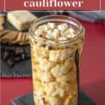 A jar of pickled cauliflower is shown with a spoon next to it. A cutting board with cheese, crackers, and dried fruits is blurred in the background. Text at the top reads "Tangy Pickled Cauliflower.