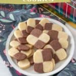 A white plate of checkerboard cookies with alternating brown and white squares is presented on a floral fabric, alongside a plaid gift box. The text at the top reads, "Checkerboard Cookies: A Classic Treat.