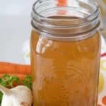 A jar of homemade vegetable stock sits on a table adorned with garlic and parsley. The text overlay reads: "Homemade Vegetable Stock - an easy & delicious recipe.