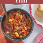 A bowl of hearty three bean chili showcasing kidney, black, and cannellini beans sits beside a dish of shredded cheese and a slice of bread, garnished with fresh herbs on the table.