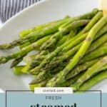 A plate of steamed asparagus with a hand squeezing a lemon wedge over it. The text reads "Fresh Steamed Asparagus.