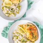 Two plates of savory lemon chicken, paired with fluffy rice and tender asparagus, rest on a pristine white surface. Garnished with fresh lemon slices, each dish is elegantly set on a backdrop of green napkins.