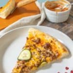 A slice of savory cheeseburger pizza rests on a plate, accompanied by a sauce dish and another tempting slice in the background.