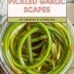 A jar of pickled garlic scapes sits on a wooden surface, the label hinting at a delightful recipe website. A soft cloth lies nearby, adding a touch of rustic charm to this delectable and tangy treat.