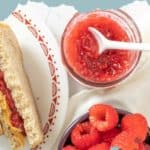 A jar of homemade raspberry jam with a spoon, fresh raspberries, and a partially visible sandwich on a plate with the text "homemade raspberry jam recipe.