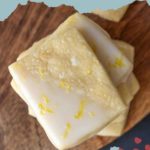 A stack of delectable lemon shortbread cookies, draped in white icing and sprinkled with zesty lemon zest, rests on a rustic wooden surface. Above them, the text "LEMON SHORTBREAD COOKIES" is elegantly framed by decorative elements.