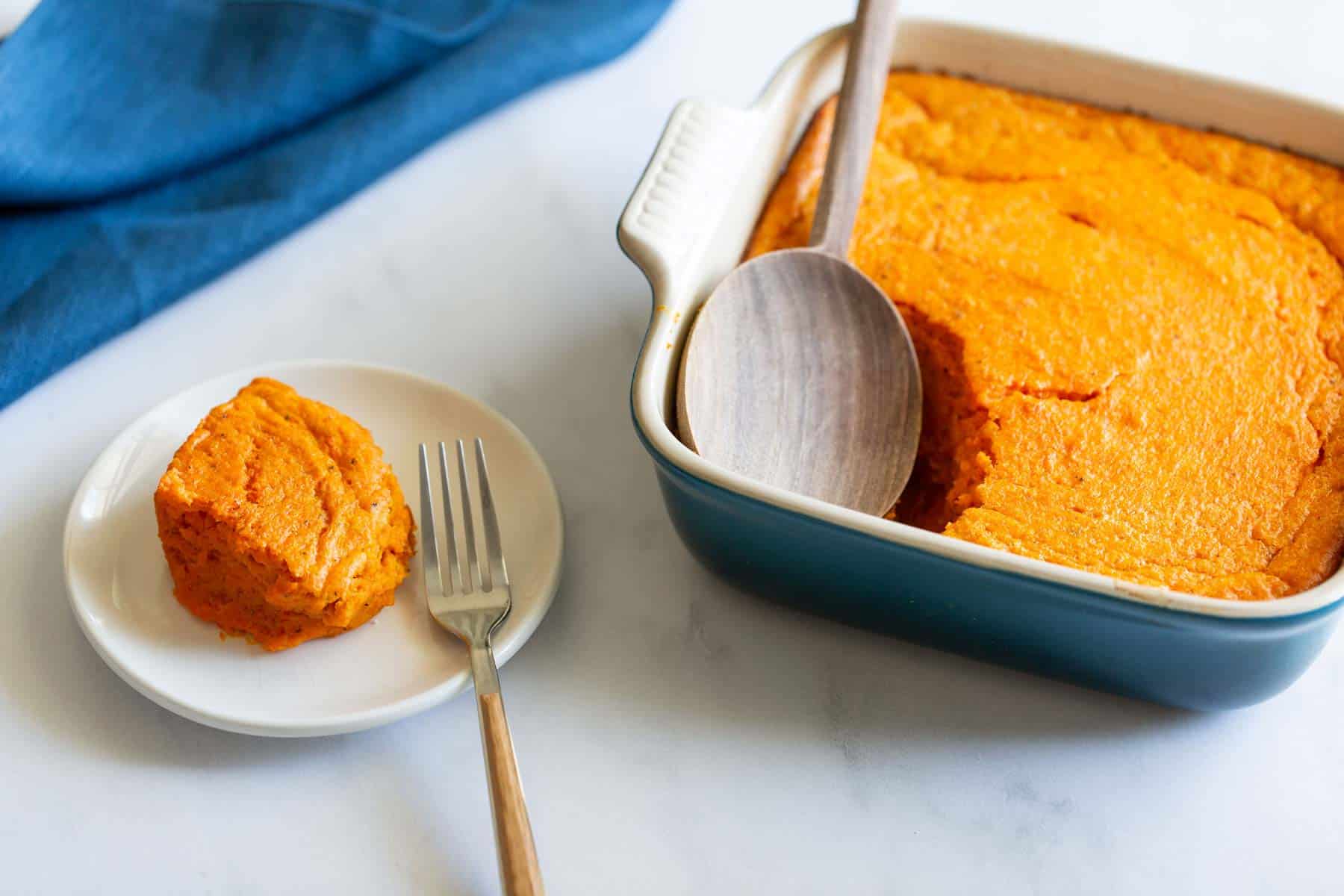 A serving of carrot souffle on a white plate with a fork, next to the baking dish.