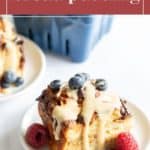 A serving of croissant bread pudding topped with blueberries and sauce, garnished with a raspberry, is on a white plate. Another plate featuring the delectable croissant bread pudding is in the background.