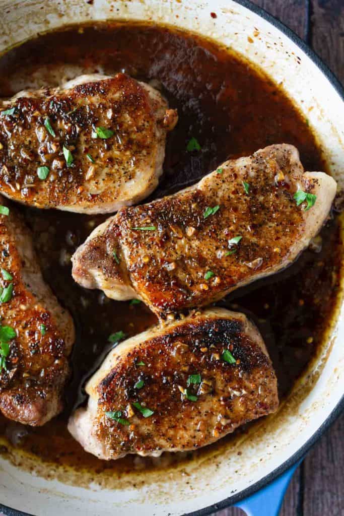 Three seared pork chops in a skillet, seasoned with herbs and spices, garnished with chopped parsley.