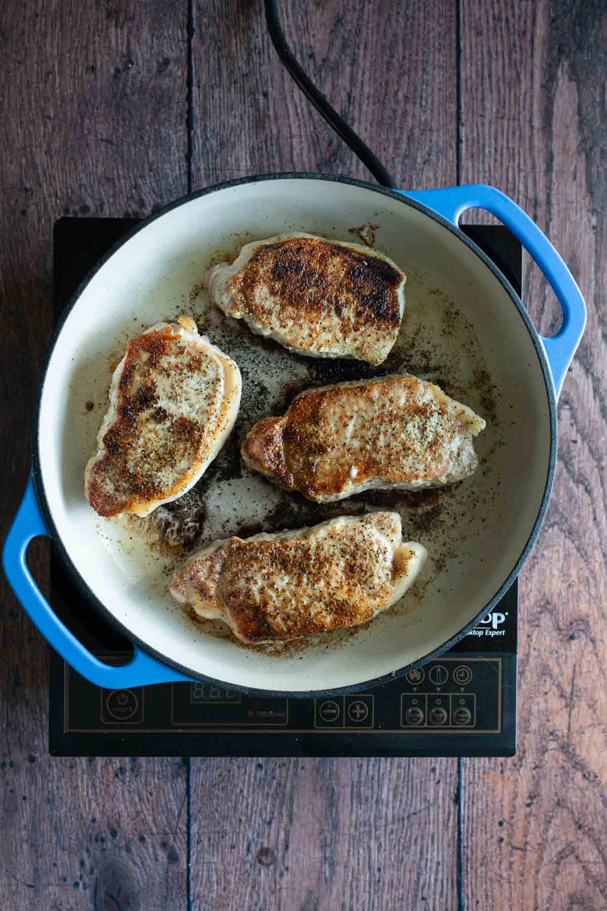 Four pork chops cooking in a white skillet on a stove, viewed from above.