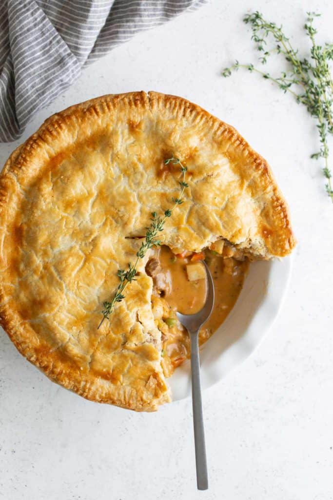 A golden beef pot pie with a slice removed, revealing its creamy filling, garnished with thyme on a white plate with a spoon.