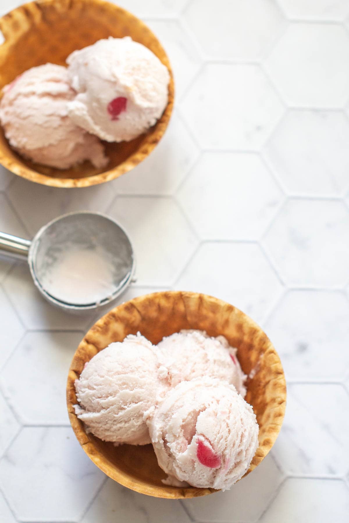 Two waffle bowls with scoops of cherry ice cream and cherry pieces, along with an ice cream scooper on a hexagon-tiled white surface.