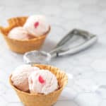 Two waffle bowls of pink and white scooped ice cream, each topped with a cherry piece. An ice cream scoop is placed between the two bowls on a marbled surface.