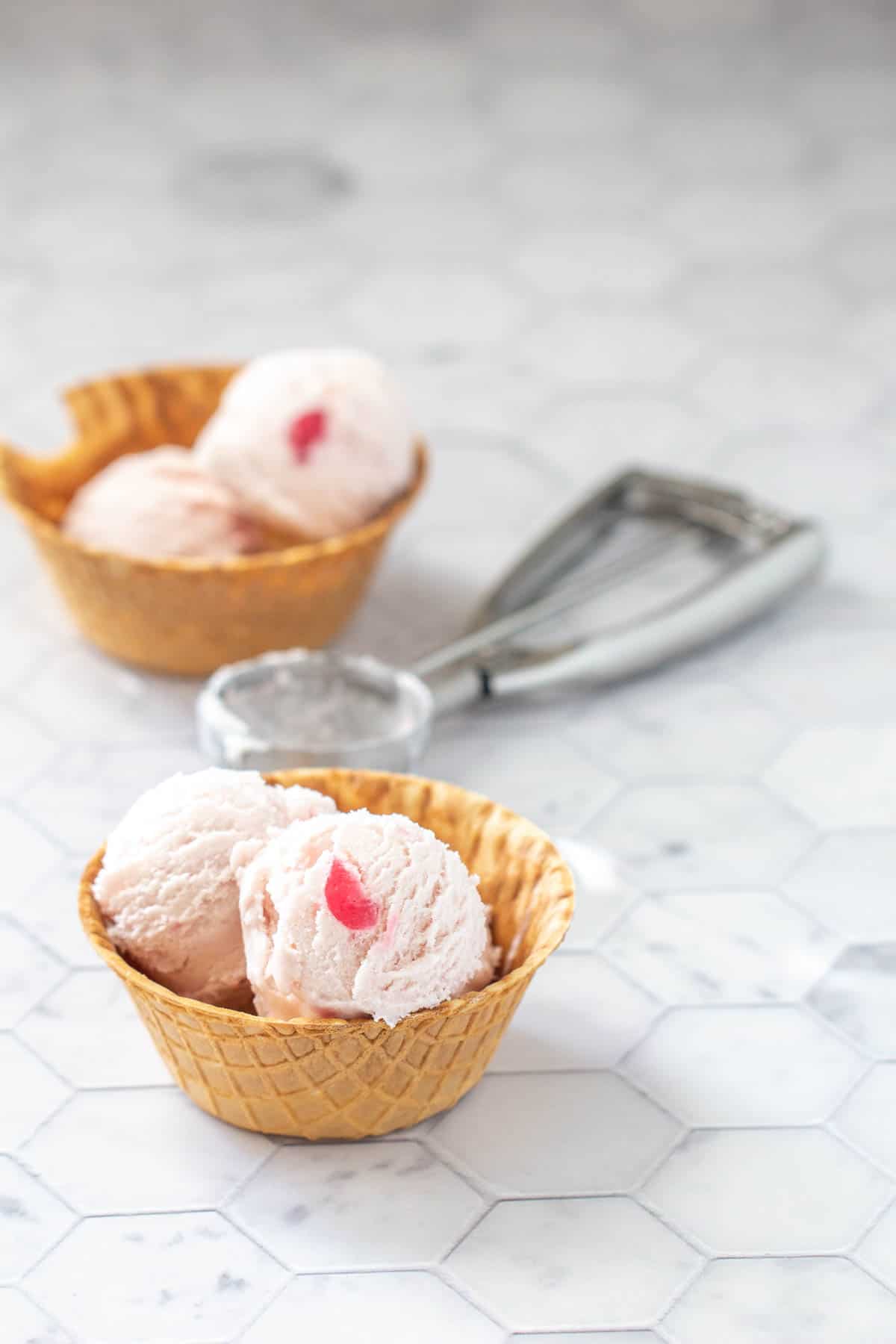 Two waffle bowls of pink and white scooped ice cream, each topped with a cherry piece. An ice cream scoop is placed between the two bowls on a marbled surface.