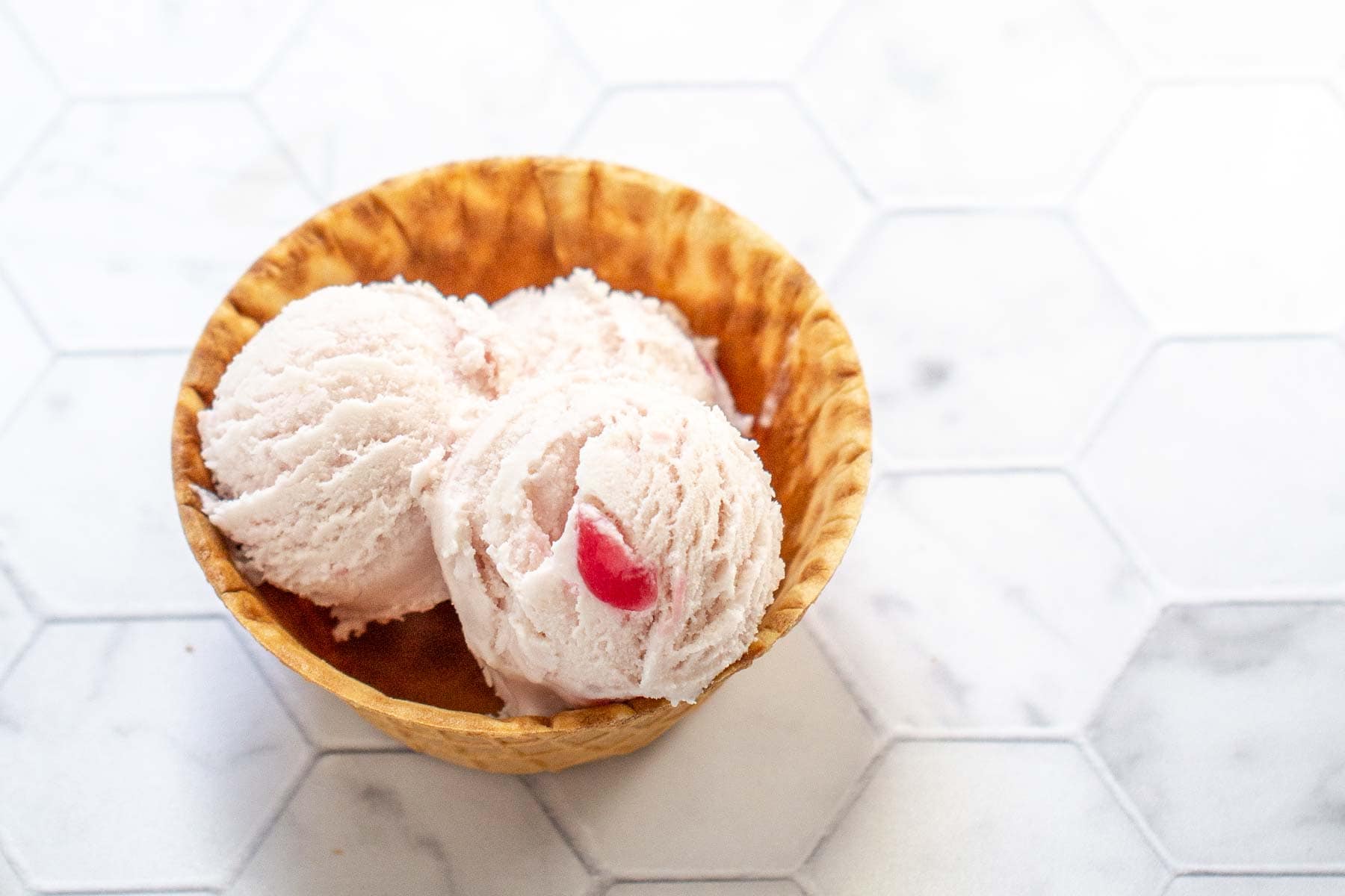 A waffle bowl contains three scoops of pink ice cream with a small red piece of fruit on top, set on a white hexagonal tiled surface.