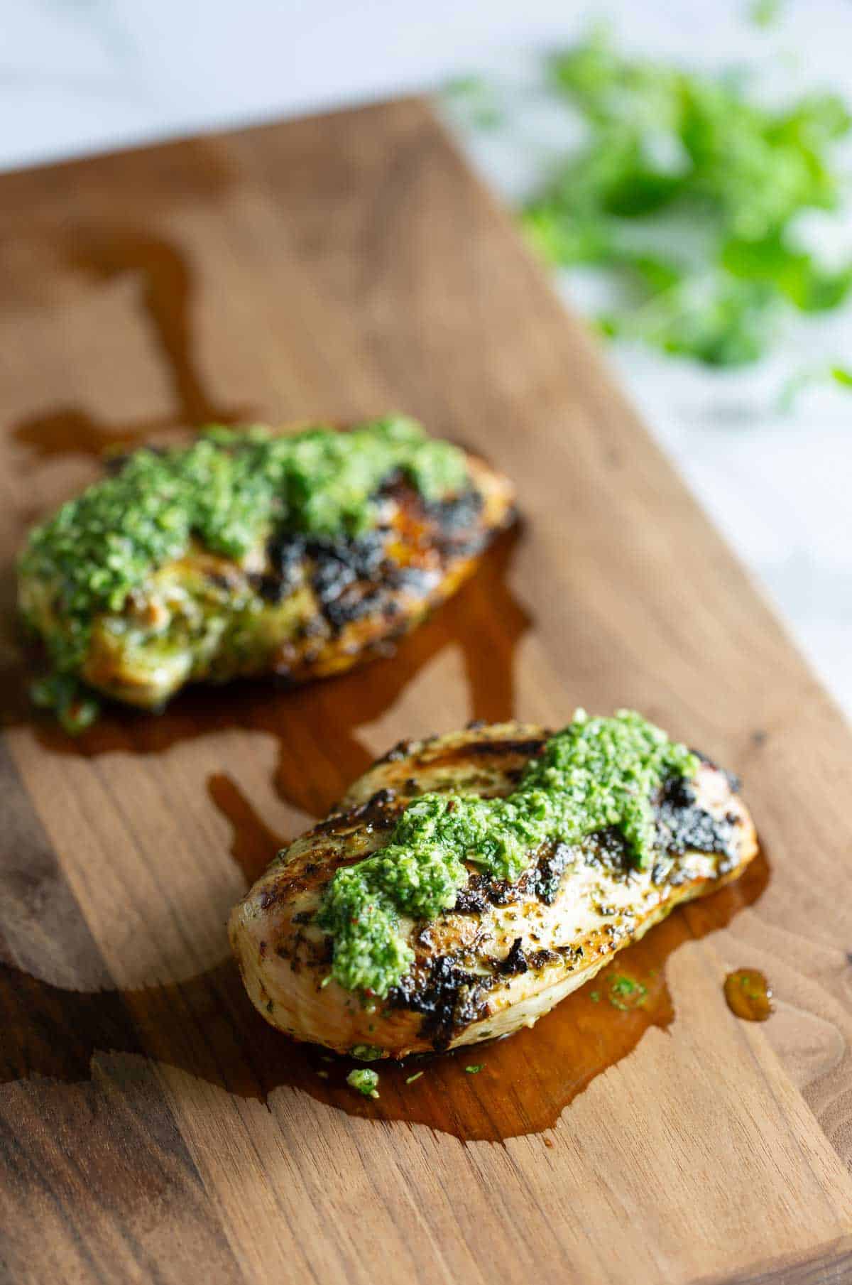 Two grilled chicken breasts topped with chimichurri sauce on a wooden cutting board, with some herbs in the background.
